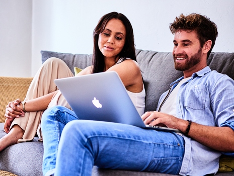 Junges Paar sitzt entspannt auf einem Sofa und schaut gemeinsam auf einen Laptop.